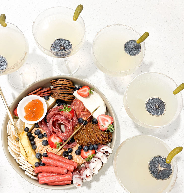 Platter of charcuterie with five cocktails garnished with pickles on a white surface